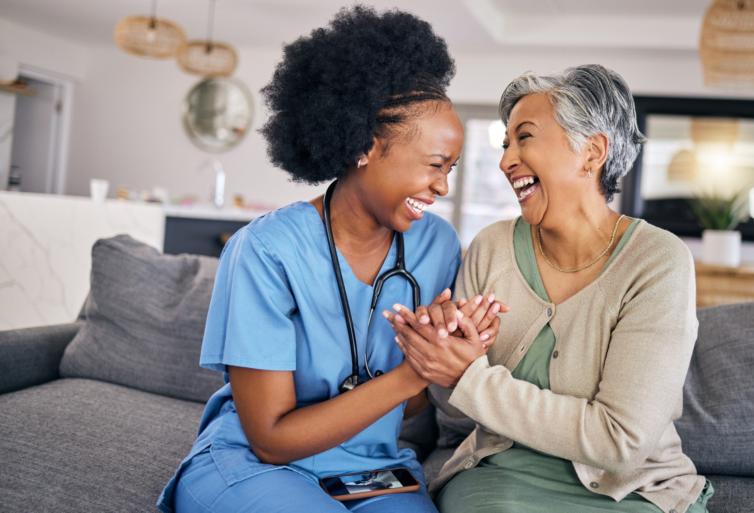 A nurse in blue scrubs and an elderly woman sit on a couch, holding hands and laughing joyfully together in a bright living room. The mood is warm, friendly, and caring.