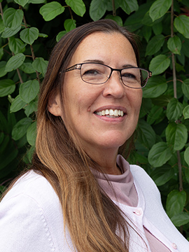 A woman with long brown hair and glasses smiles, wearing a light pink top and white sweater, standing in front of green leafy plants.