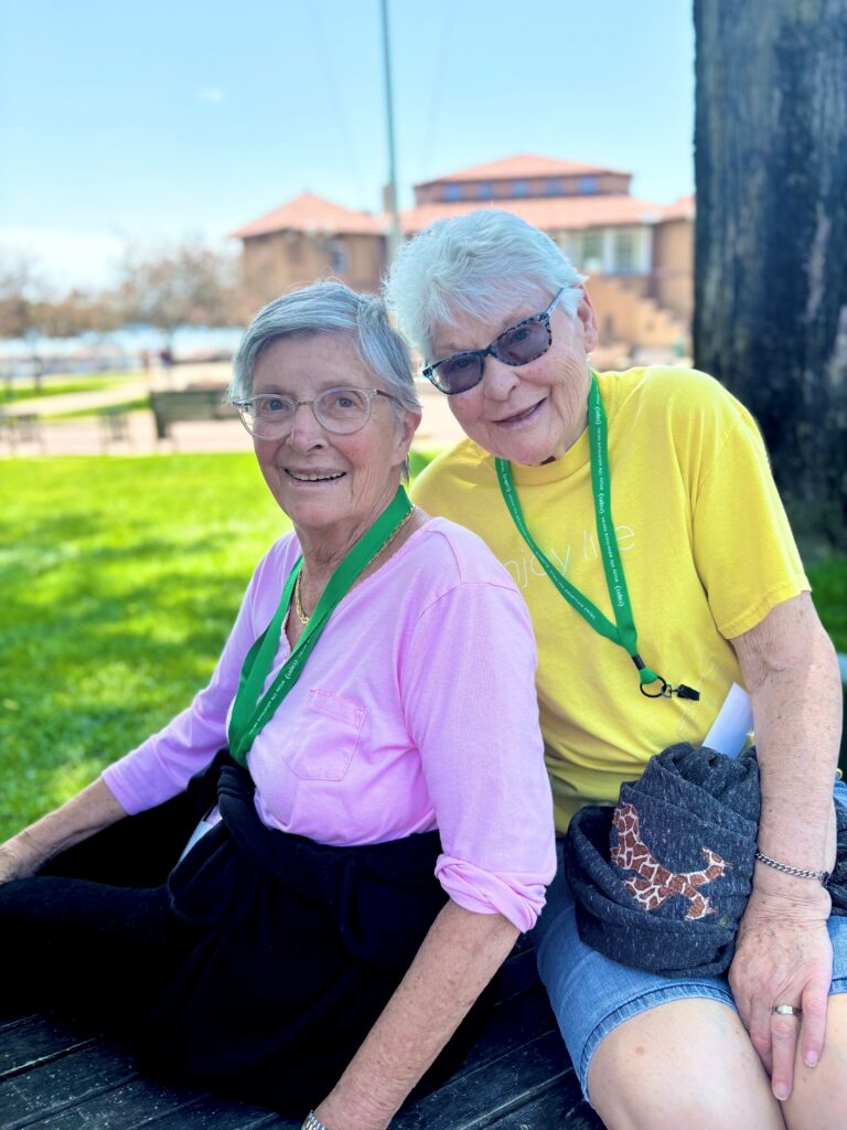 Two older women sit together on a bench outside, smiling. One wears a pink shirt, glasses, and a green lanyard; the other wears a yellow shirt, sunglasses, and a waist pouch. Bright, sunny day in a park.
