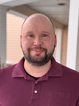 A man with a shaved head and a short beard, wearing a maroon polo shirt, stands outside under a covered walkway with beige siding in the background.