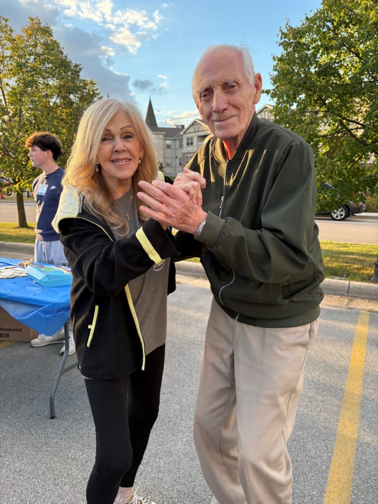 An elderly man and woman smile while dancing together outdoors in a parking lot, with trees, a table, and a building visible in the background on a sunny day.