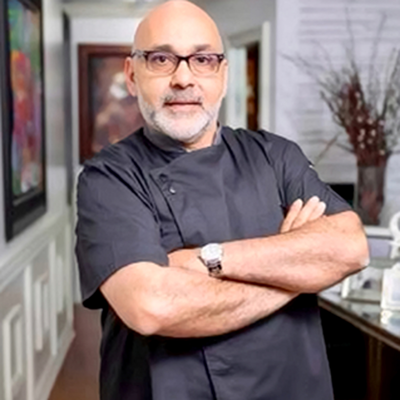 A man with glasses and a beard, wearing a dark chefs coat, stands with arms crossed in a modern, well-lit room decorated with art and a vase of branches in the background.
