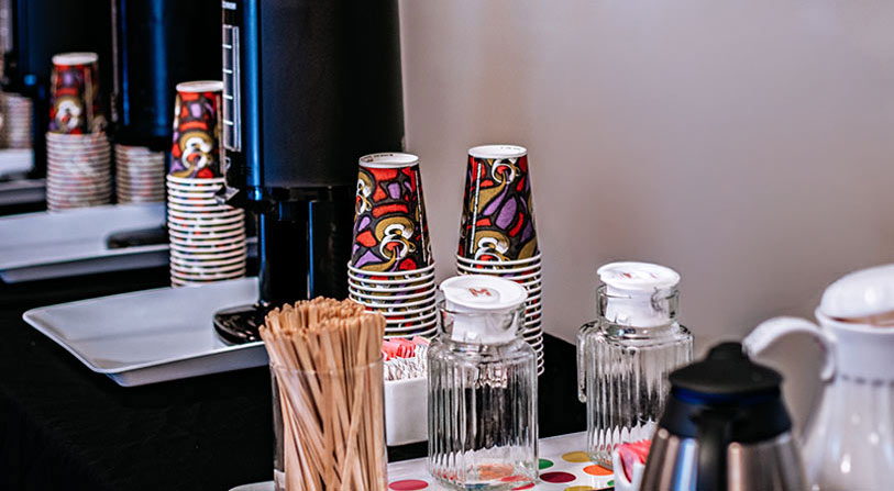 A coffee station with stacked colorful paper cups, large coffee dispensers, glass jars of sugar and creamer, wooden stir sticks, and a tray with packets of sweetener on a table.
