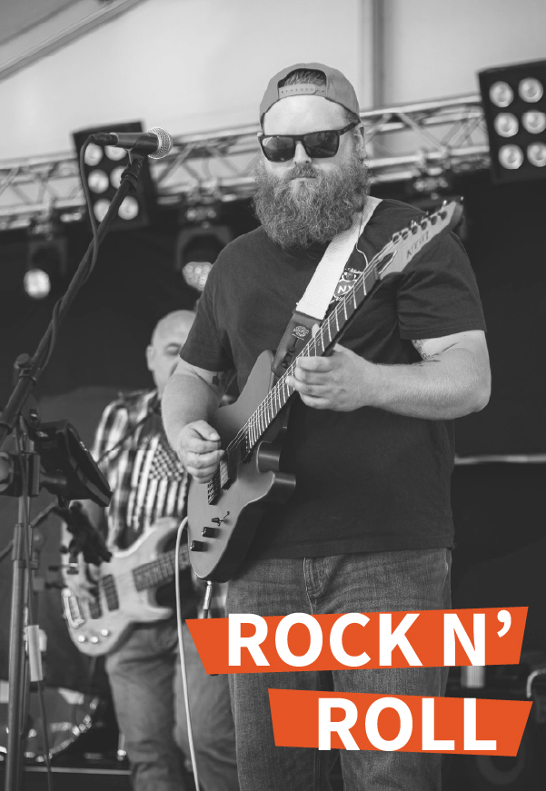 A bearded musician wearing sunglasses and a cap plays electric guitar on stage, with a bassist in the background. Bright stage lights shine above. The text ROCK N’ ROLL is displayed in bold letters at the bottom right.