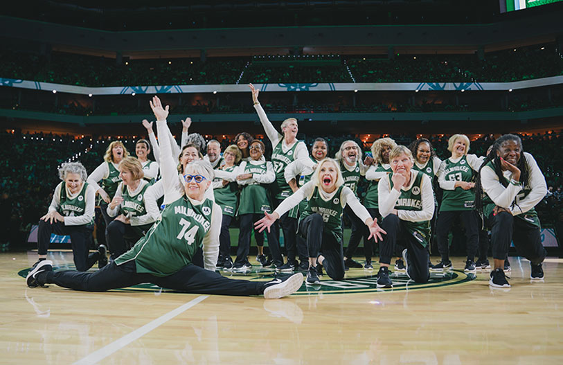 A group of enthusiastic older women in green basketball jerseys pose energetically on a basketball court, with one woman doing the splits and others smiling and cheering in a stadium setting.