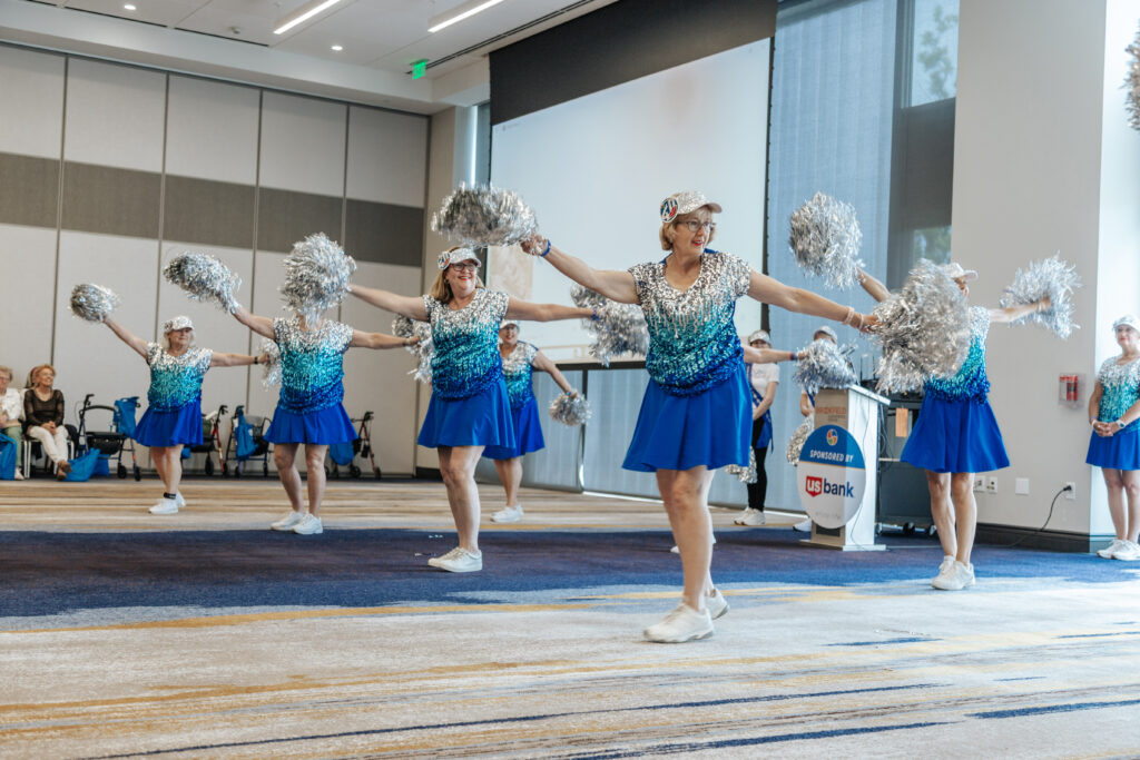 A group of older women in matching blue skirts, white sneakers, and silver pom-poms perform a cheerleading routine indoors, smiling and extending their arms, with an audience seated in the background.
