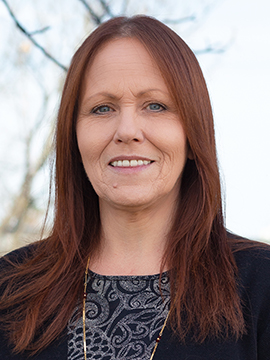 A woman with straight, reddish-brown hair and light skin is standing outdoors, wearing a dark patterned top and a black sweater. Leafless tree branches and a bright sky are visible in the background.
