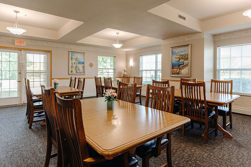A bright dining room with wooden tables and chairs, flower centerpieces, large windows letting in natural light, artwork on the walls, and a door leading outside.