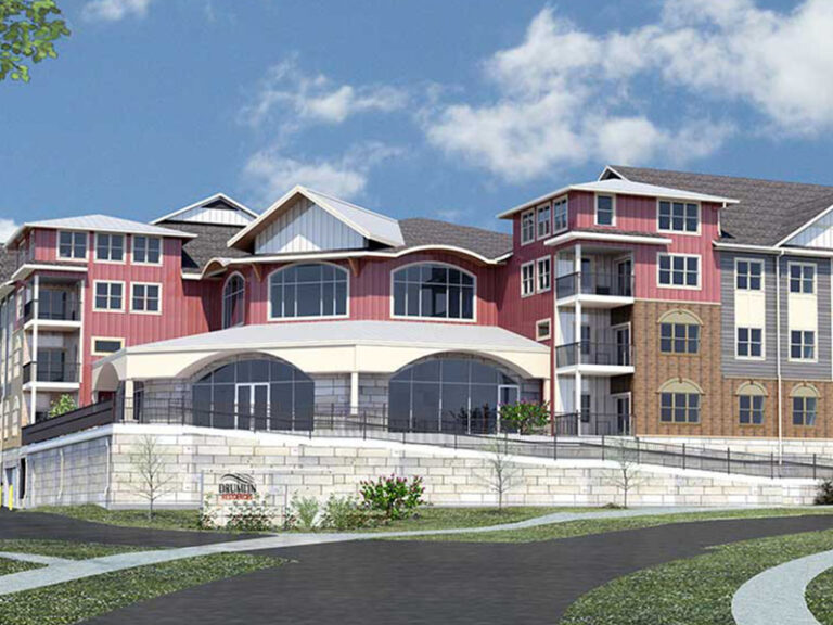 A modern multi-story building with red and beige siding, large windows, and balconies, set against a partly cloudy sky. A sign and landscaping are visible at the front of the building.