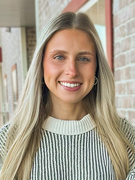 A young woman with long blonde hair smiles at the camera. She is wearing a white and black striped sweater and standing in front of a brick building with windows.