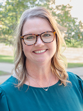A woman with wavy blonde hair and glasses smiles outdoors, wearing a teal blouse and a silver necklace. Green trees and a blurred pathway are visible in the background.