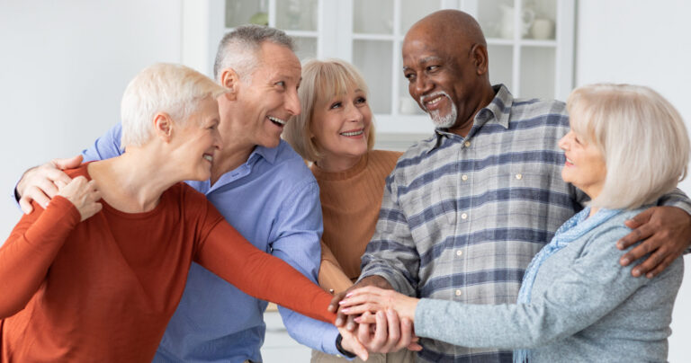 Five smiling older adults stand together indoors, placing their hands in a pile in the center, showing unity and friendship. They are casually dressed and appear happy and engaged with each other.