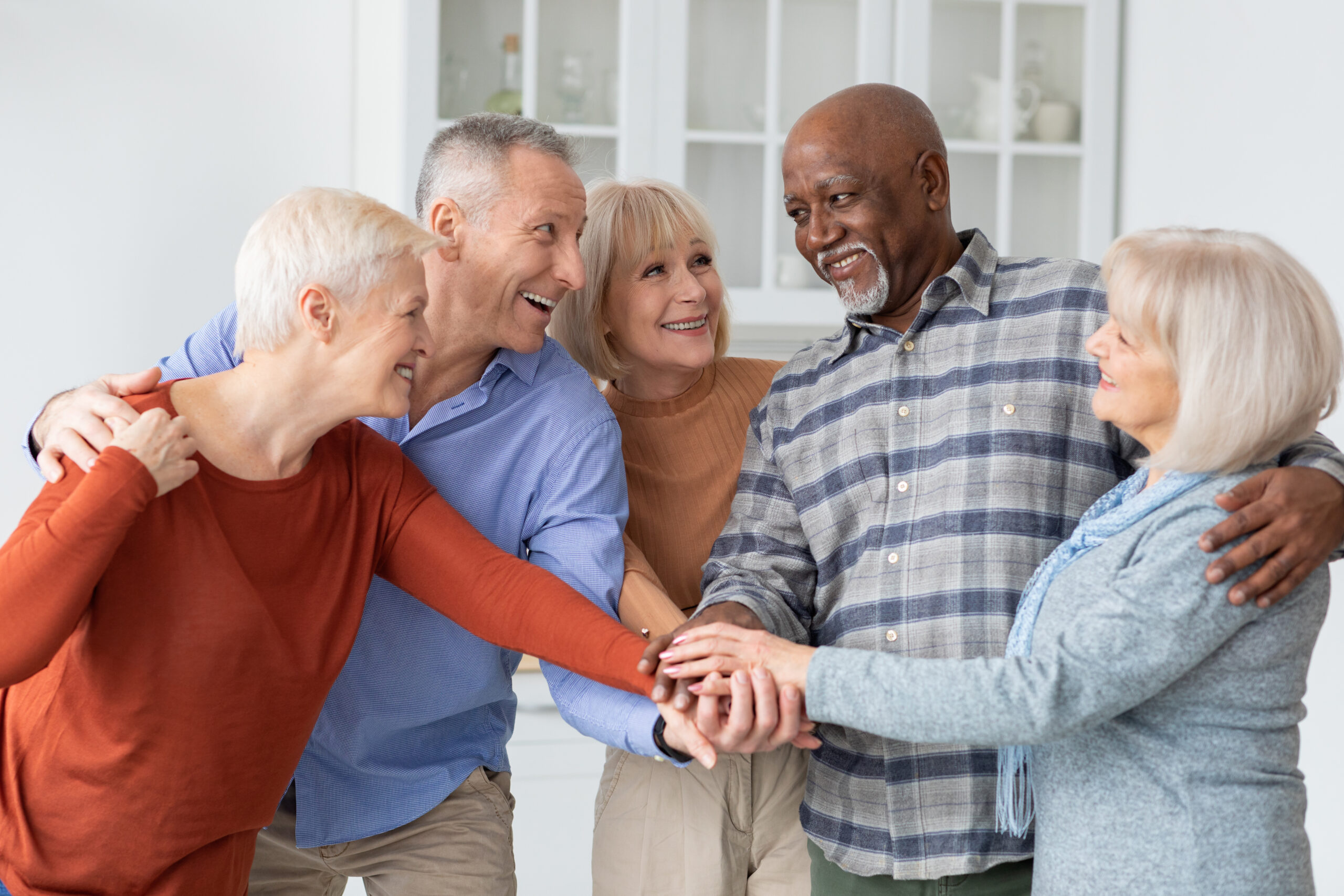 Five smiling older adults stand together indoors, placing their hands in a pile in the center, showing unity and friendship. They are casually dressed and appear happy and engaged with each other.