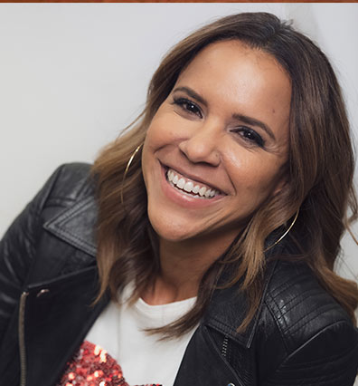 A woman with brown hair, wearing a black leather jacket and hoop earrings, smiles broadly at the camera against a light background.