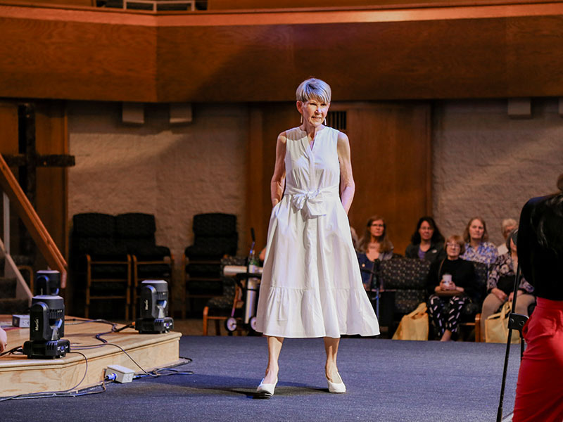 A woman in a sleeveless white dress and white shoes stands confidently on a stage, posing for an audience seated in the background. The setting appears to be an indoor event or fashion show.
