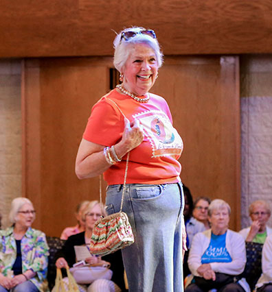 An older woman with gray hair, wearing a red t-shirt, denim skirt, and beaded accessories, smiles while walking on a runway. She carries a small purse and an audience sits in the background.