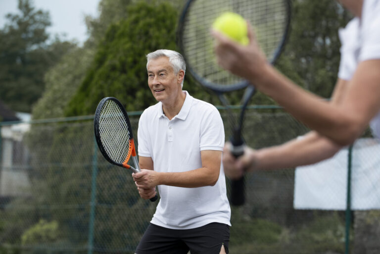 An older man in a white polo shirt prepares to play tennis, holding a racket and smiling, while another player in the foreground holds a tennis ball and racket. They are outdoors on a court with trees in the background.