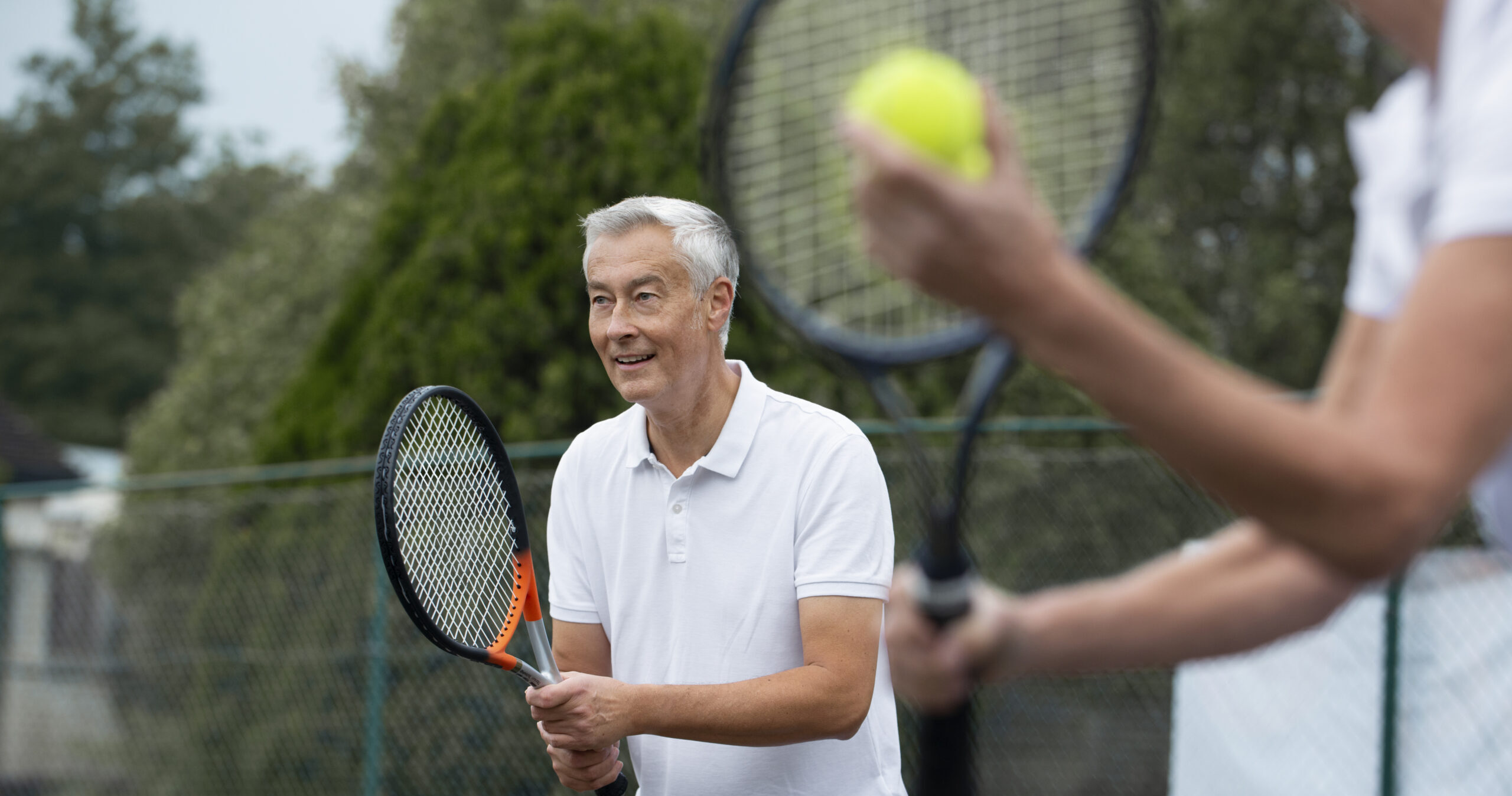 An older man in a white polo shirt prepares to play tennis, holding a racket and smiling, while another player in the foreground holds a tennis ball and racket. They are outdoors on a court with trees in the background.