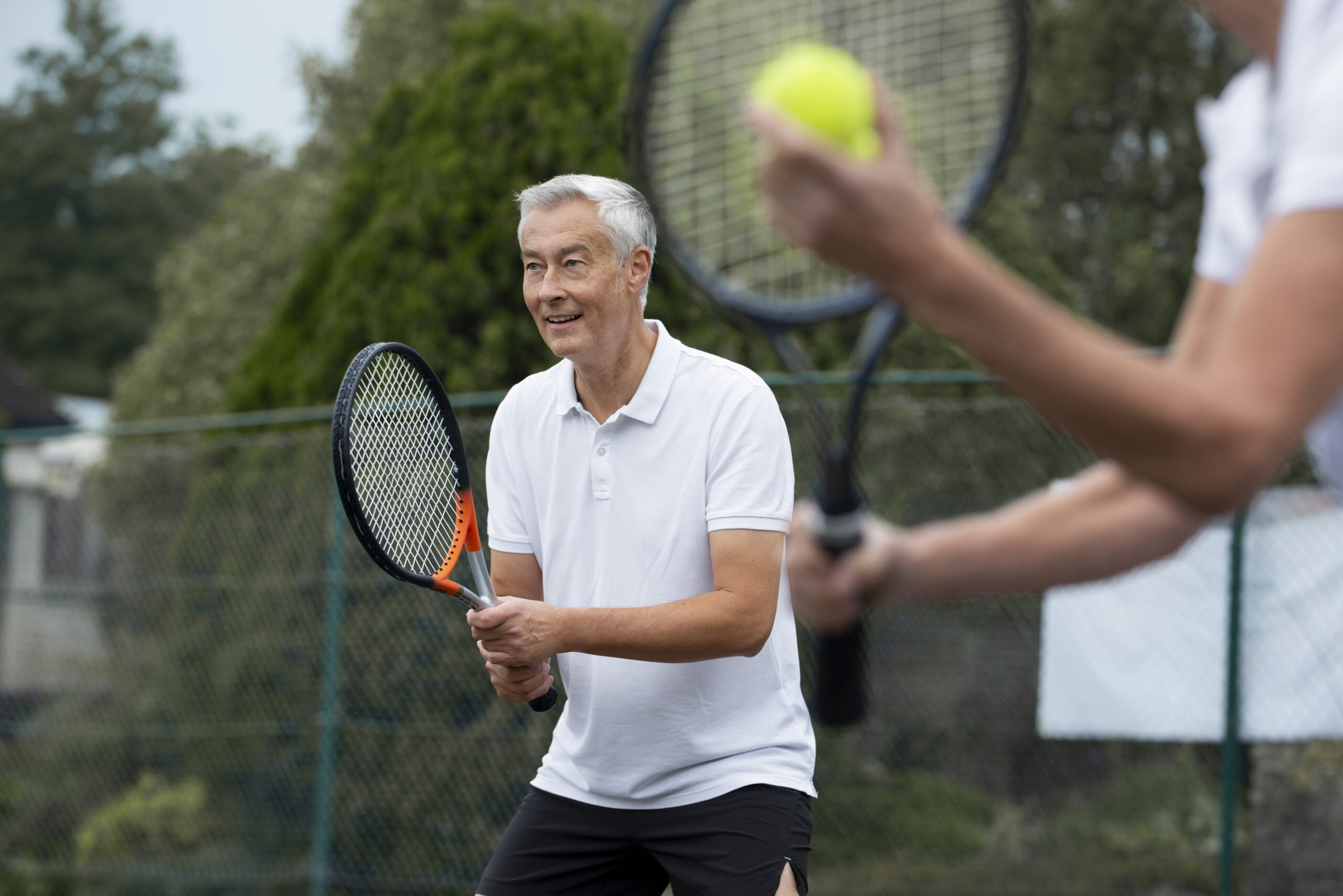 An older man in a white polo shirt prepares to play tennis, holding a racket and smiling, while another player in the foreground holds a tennis ball and racket. They are outdoors on a court with trees in the background.