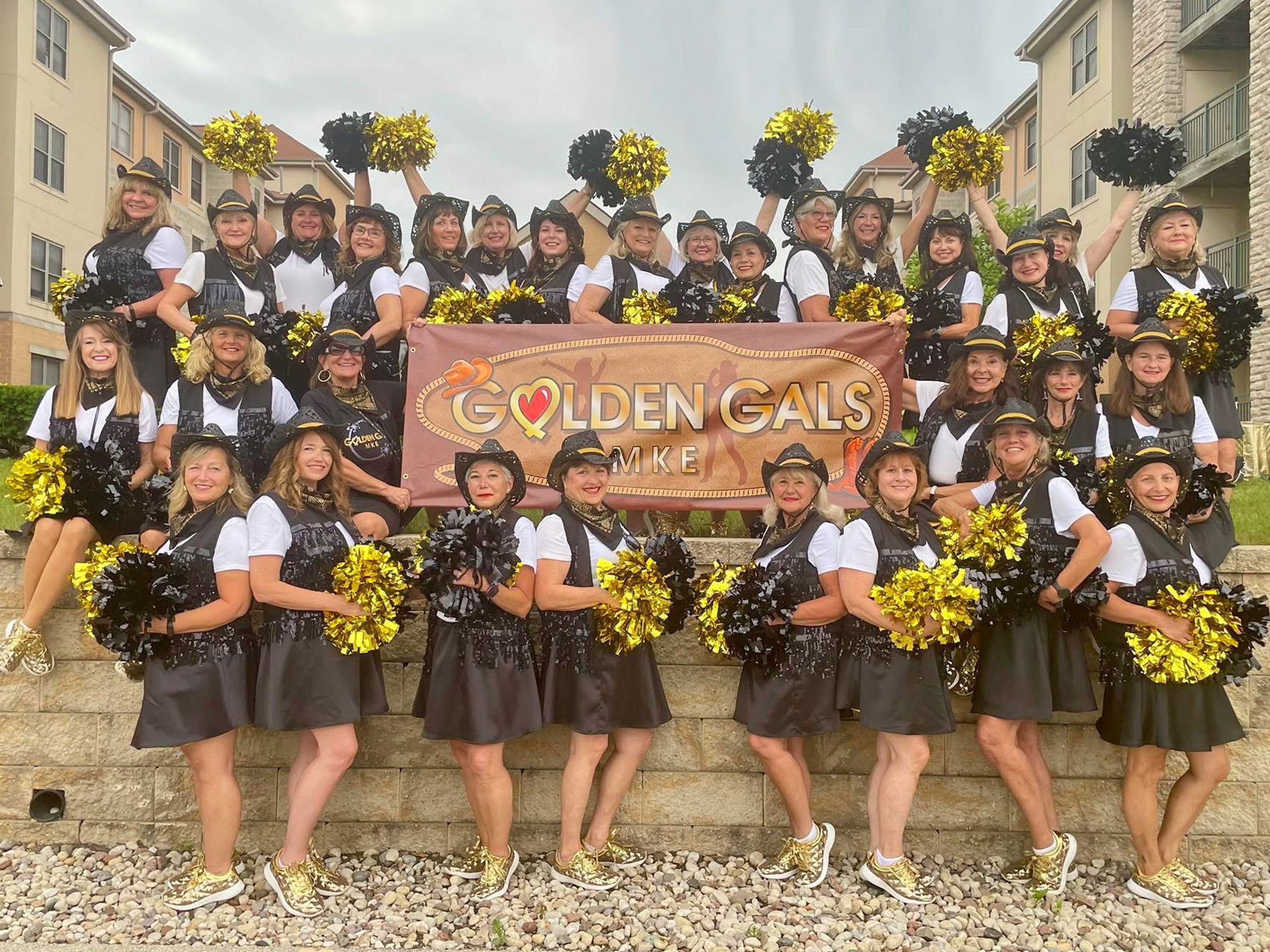 A large group of women in matching black outfits and gold shoes pose outdoors, holding gold and black pom-poms. They stand behind a banner that reads Golden Gals. Apartment buildings are visible in the background.