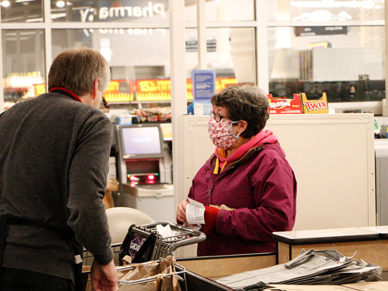 A cashier wearing a dark sweater speaks to a customer in a pink mask and maroon jacket at a supermarket checkout counter. Shopping bags and candy bars are visible on the counter.