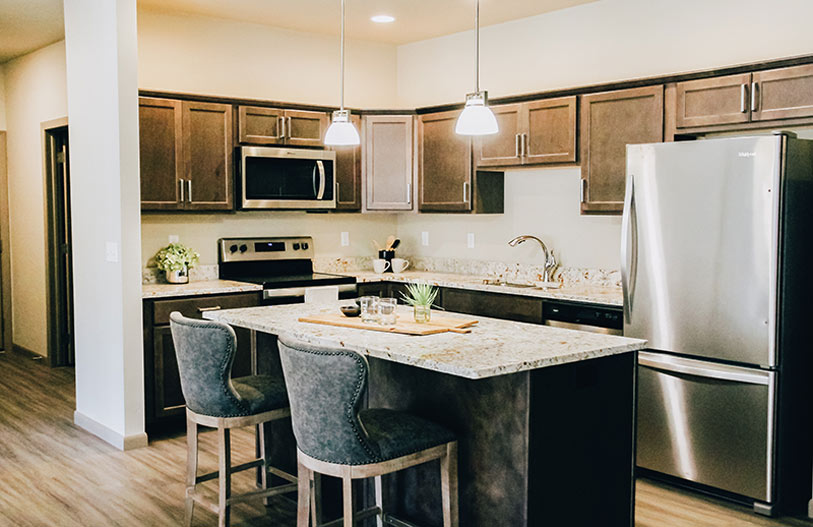 Modern kitchen with brown cabinets, stainless steel appliances, granite countertops, an island with two gray upholstered chairs, hanging pendant lights, and decorative plants.
