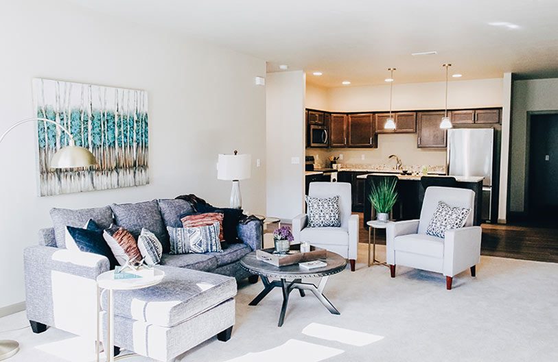 Bright, modern open-concept living room and kitchen featuring a gray sofa, two light armchairs, a round coffee table, decorative pillows, wall art, and pendant lighting over dark wood kitchen cabinets.