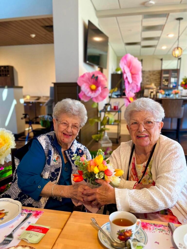 Two elderly women sitting at a table in a brightly decorated room, holding a colorful bouquet of flowers and smiling. Large pink paper flowers and tea cups are on the table.