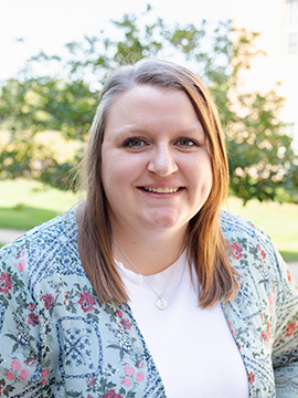 A woman with straight, light brown hair wearing a white shirt and a blue floral-patterned cardigan smiles outdoors with green trees and sunlight in the background.