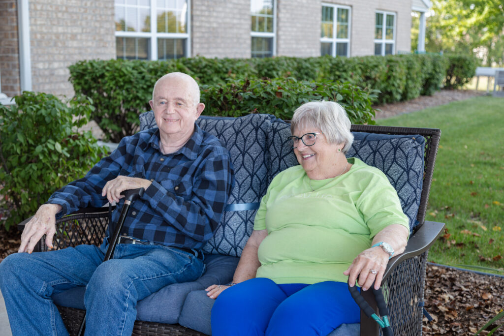 An elderly man and woman sit together on an outdoor bench, smiling and enjoying a sunny day. They are in front of a brick building with green bushes and windows in the background.