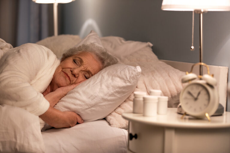 An elderly woman with gray hair sleeps peacefully in bed, resting her head on a white pillow. A bedside table holds an alarm clock, a lamp, and several medicine bottles. The room has a calm, cozy atmosphere.