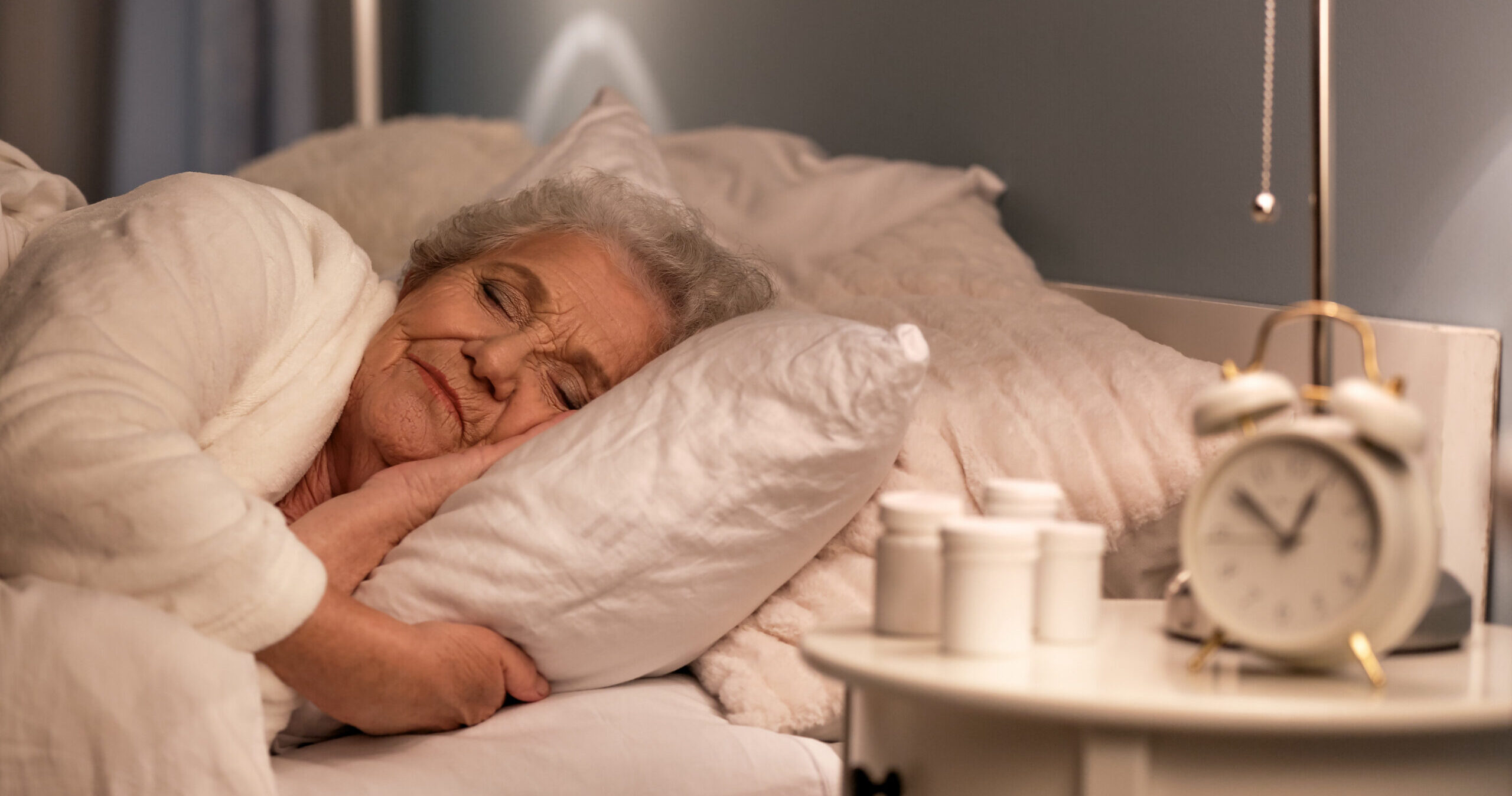 An elderly woman with gray hair sleeps peacefully in bed, resting her head on a white pillow. A bedside table holds an alarm clock, a lamp, and several medicine bottles. The room has a calm, cozy atmosphere.