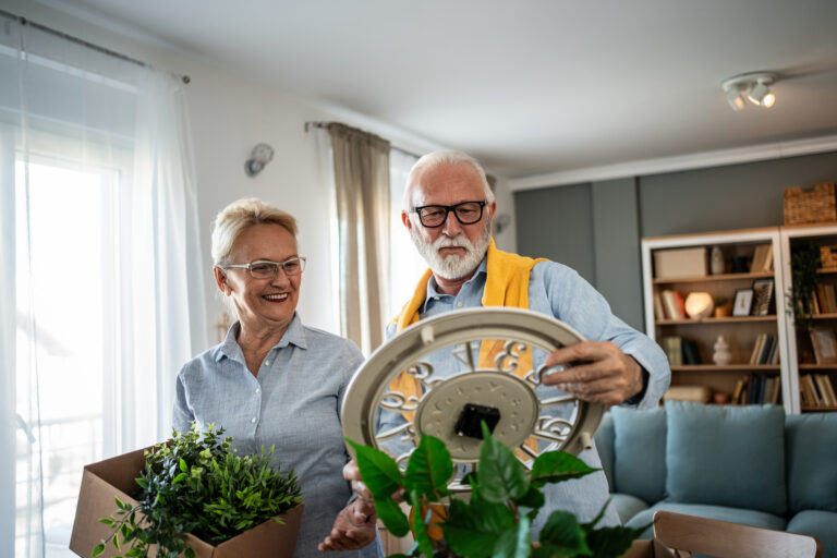 An older man and woman stand in a bright living room. The woman holds a box with a potted plant, smiling at the man, who examines a round wall clock among more plants. Shelves with books are in the background.