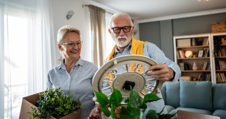 An older man and woman stand in a bright living room. The woman holds a box with a potted plant, smiling at the man, who examines a round wall clock among more plants. Shelves with books are in the background.