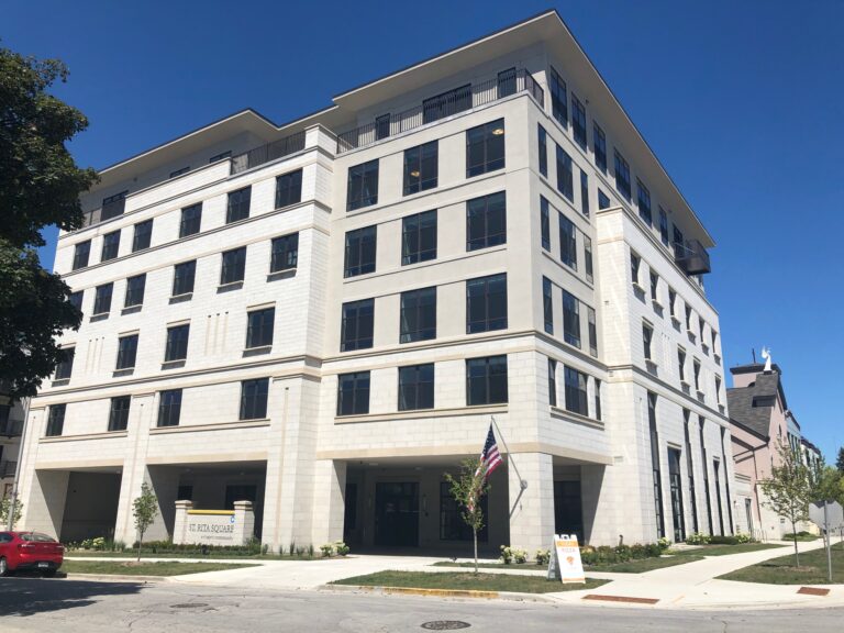 A modern, multi-story beige building with large windows, a U.S. flag at the entrance, a red car parked nearby, and a clear blue sky in the background. A sign is placed on the grass near the sidewalk.