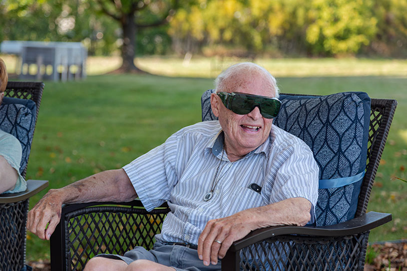 An elderly man wearing dark sunglasses sits comfortably in a patio chair outdoors, smiling. He wears a striped short-sleeve shirt and shorts, with trees and grass visible in the background.