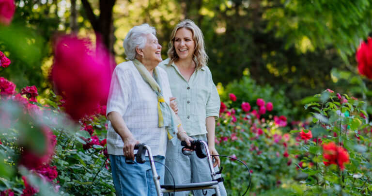 An elderly woman with a walker and a younger woman smile together while standing in a garden filled with vibrant red flowers and lush greenery.