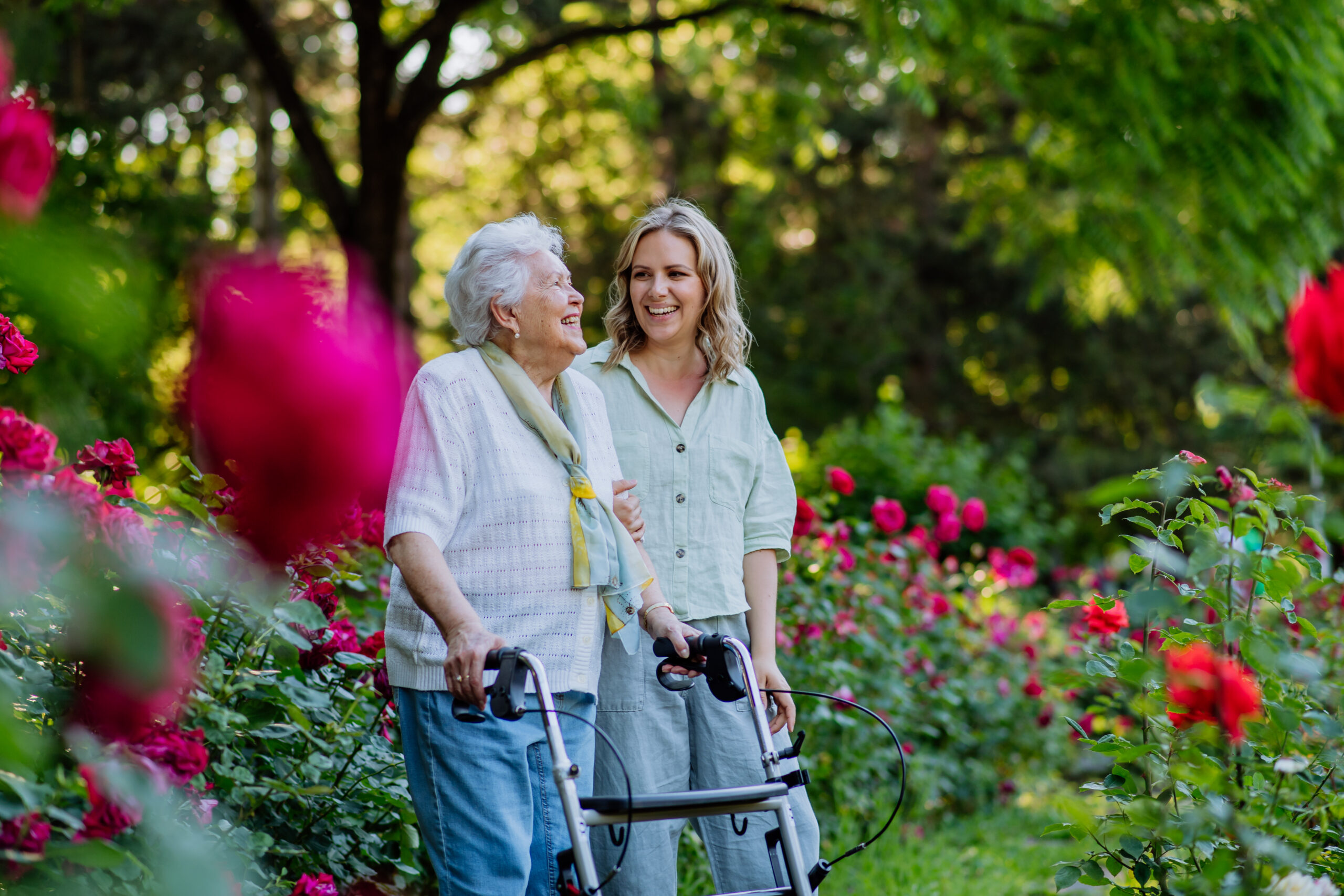 An elderly woman with a walker and a younger woman smile together while standing in a garden filled with vibrant red flowers and lush greenery.