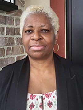 A woman with short, curly, light blonde hair stands in front of a brick wall. She is wearing hoop earrings, a black blazer, and a white top with a red and green pattern. She has a neutral expression.