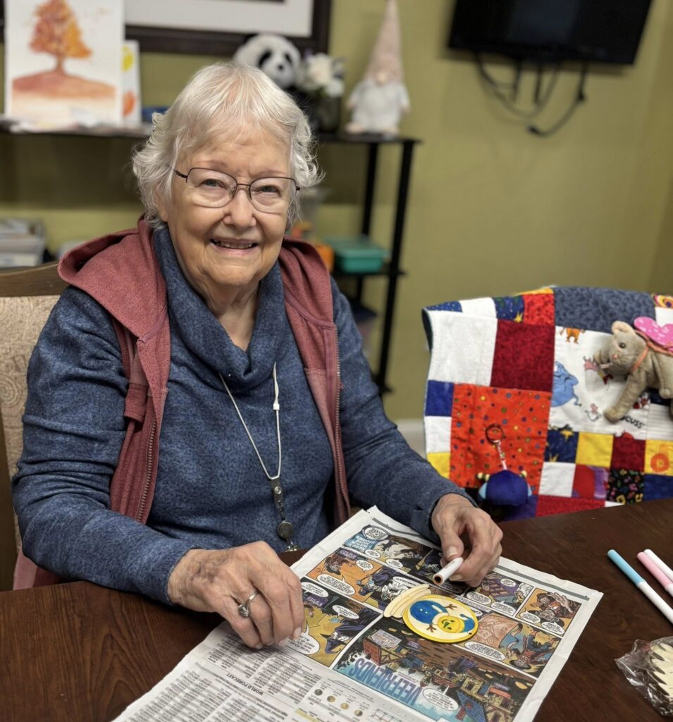 An elderly woman with white hair and glasses smiles while sitting at a table coloring a newspaper comic. She wears a blue hoodie and gray vest. A colorful quilt and stuffed elephant are on a nearby chair.