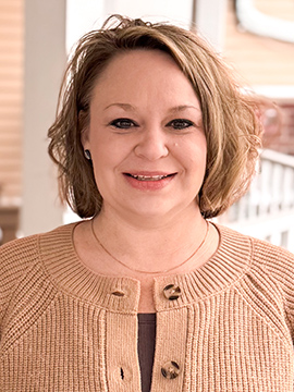 A woman with short, wavy light brown hair smiles at the camera. She is wearing a tan knit sweater with large buttons and a dark top underneath, standing in front of a porch railing and blurred background.