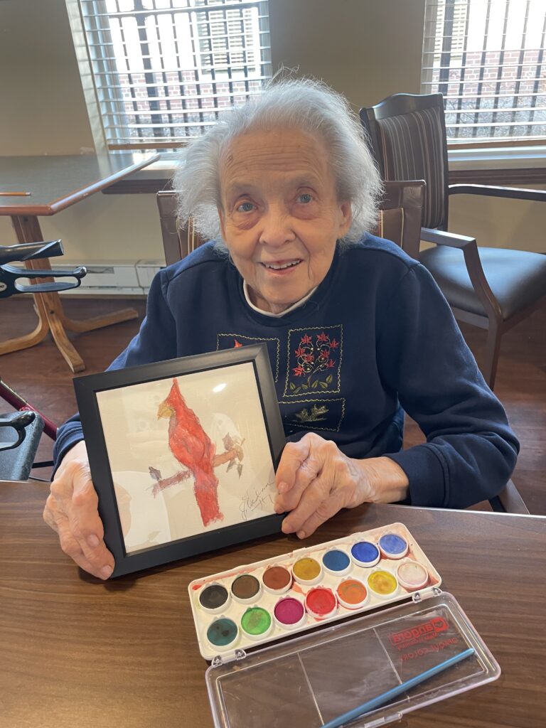 An older woman with gray hair smiles while holding a framed watercolor painting of a red cardinal. In front of her on the table are watercolor paints and a brush. Sunlight streams through window blinds behind her.