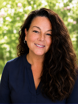 A woman with long, curly brown hair wearing a navy blue blouse is smiling at the camera. The background is blurred greenery with soft light.