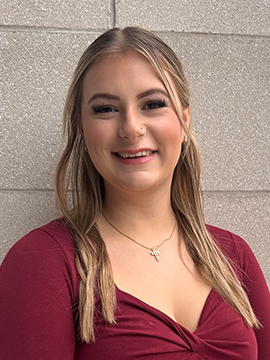 A young woman with long, light brown hair is smiling in front of a light gray brick wall. She is wearing a maroon top and a gold necklace with a small pendant.
