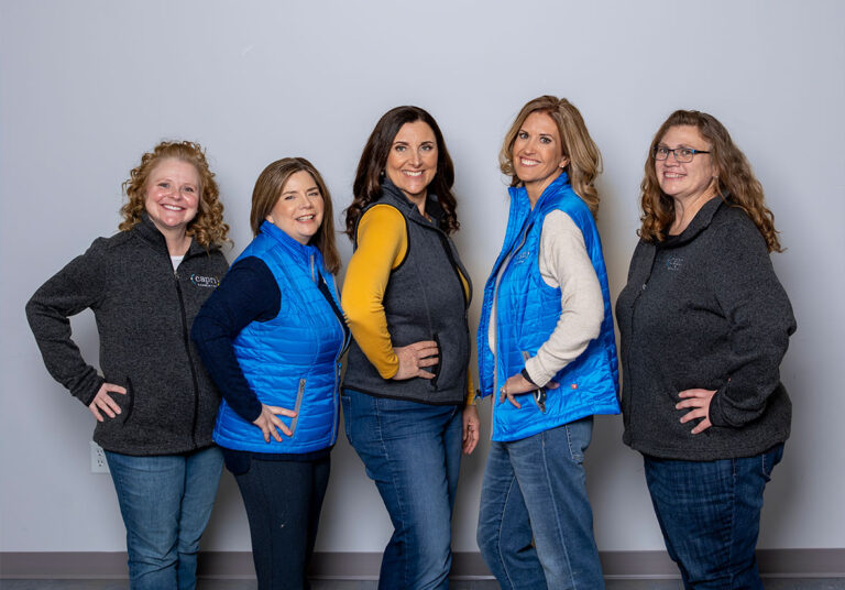Five women stand side by side, smiling at the camera. Three wear blue vests over sweaters and two wear dark gray zip-up sweaters. They pose with hands on hips against a plain light gray wall.
