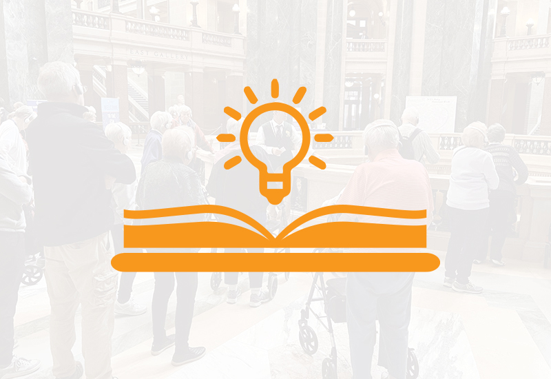 A group of people gathers in a large, ornate building; overlaid is an orange icon of an open book with a glowing light bulb above it, symbolizing learning or knowledge.