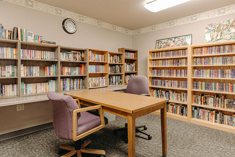 A quiet library room with wooden bookshelves filled with books, a wooden table with two purple chairs, a wall clock, and a framed map on the wall. The room is well-lit and has carpeted flooring.