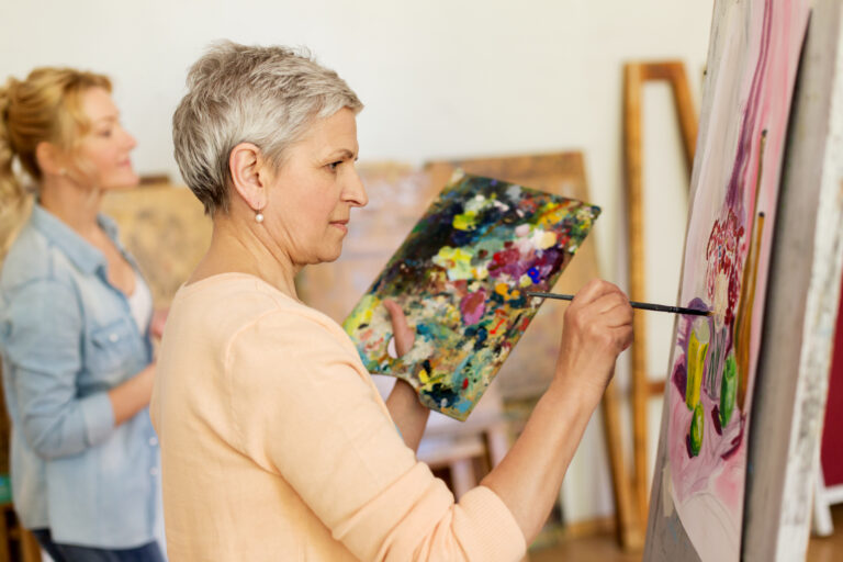An older woman with short gray hair holds a colorful palette and paints on a canvas. Another woman in the background is also painting. Both are in a bright art studio setting.