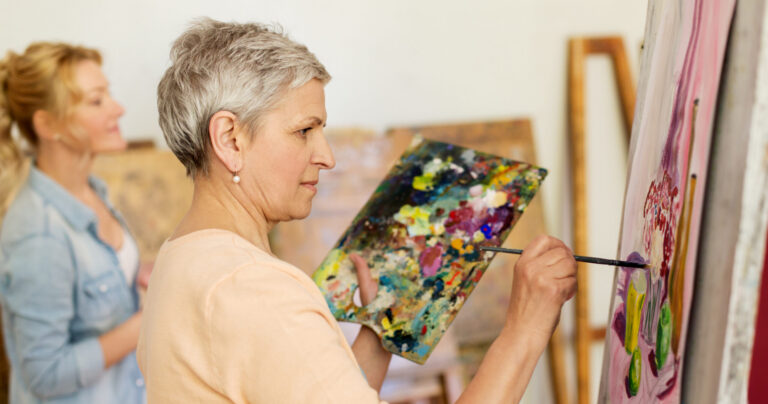 An older woman with short gray hair holds a colorful palette and paints on a canvas. Another woman in the background is also painting. Both are in a bright art studio setting.