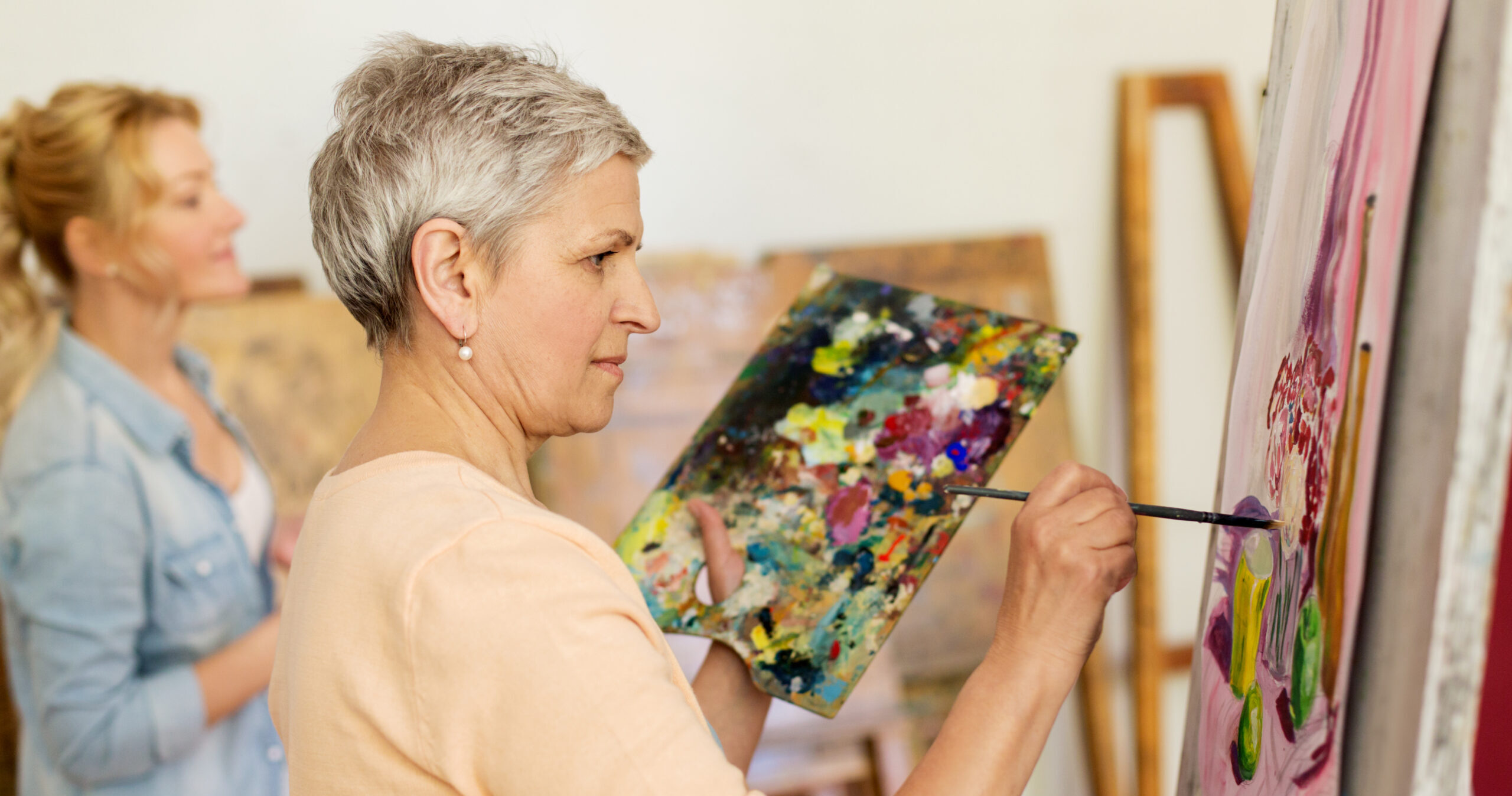 An older woman with short gray hair holds a colorful palette and paints on a canvas. Another woman in the background is also painting. Both are in a bright art studio setting.
